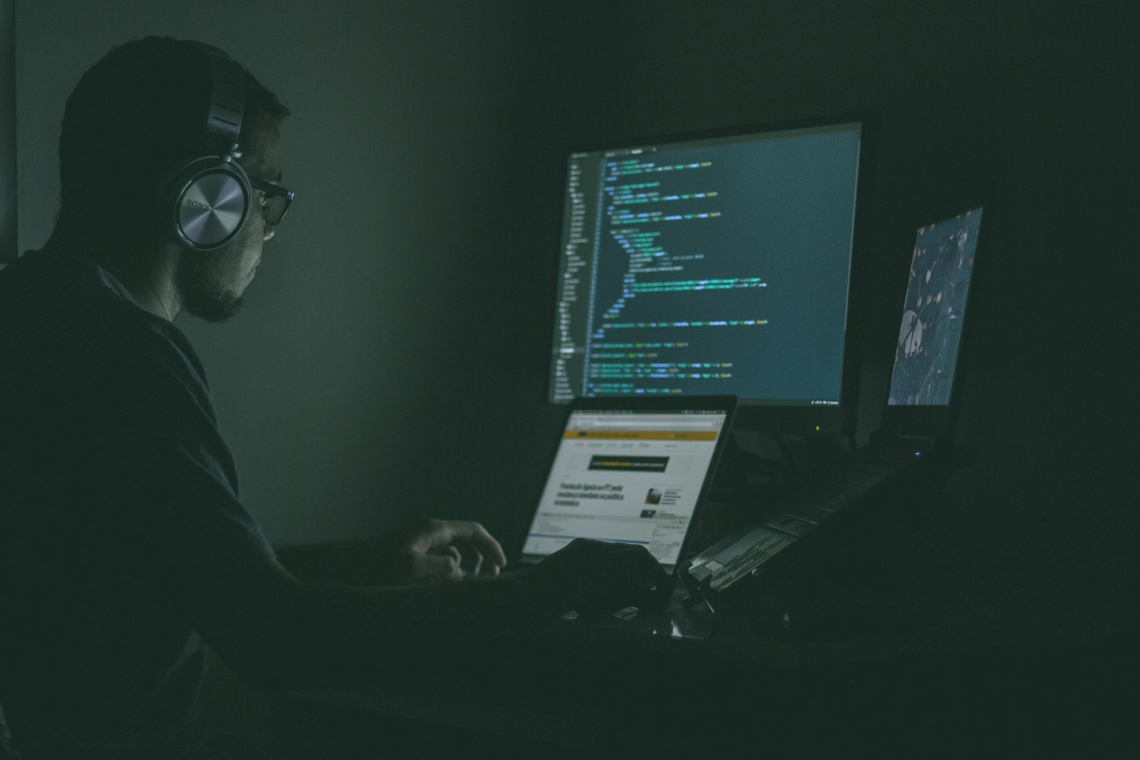 A developer wearing headphones writes code across a laptop and monitor in a softly lit workspace.